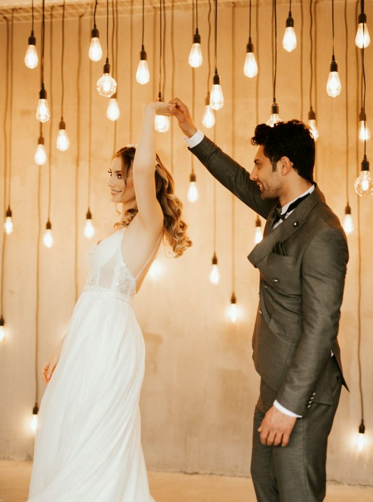 a-bride-and-groom-standing-in-front-of-a-string-of-lights-zcge3ckz7fi